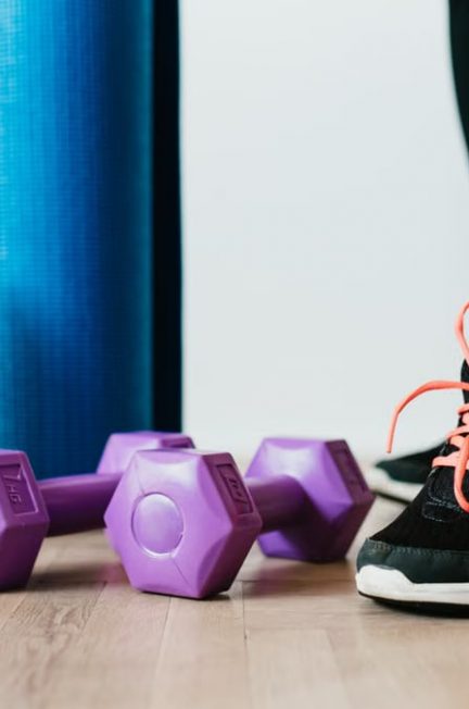 Crop sportswoman wearing sneakers and black leggings standing with sport mat on wooden floor near dumbbells and water bottle before exercising indoors