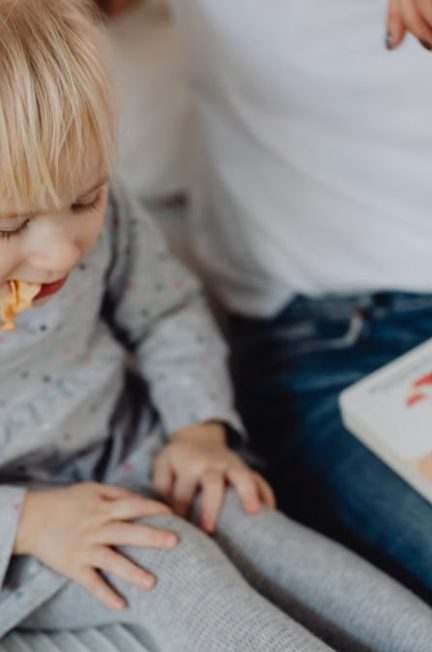 Cute toddler with ponytails enjoying a snack next to a book and parent. Cozy indoor scene.